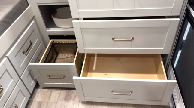 Travel trailer kitchen storage with multiple open drawers, including deep drawers and a pull-out basket organizer next to cabinetry and appliances.