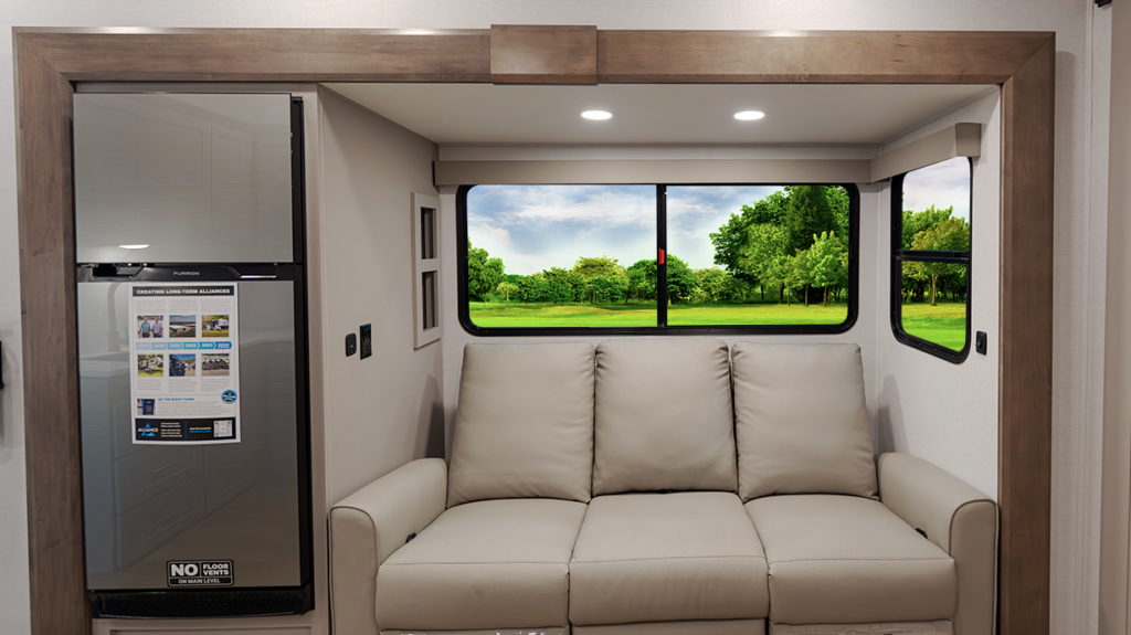 Travel trailer rear seating area with a three-cushion sofa centered beneath large panoramic windows, with a refrigerator to the left and scenic outdoor view visible.