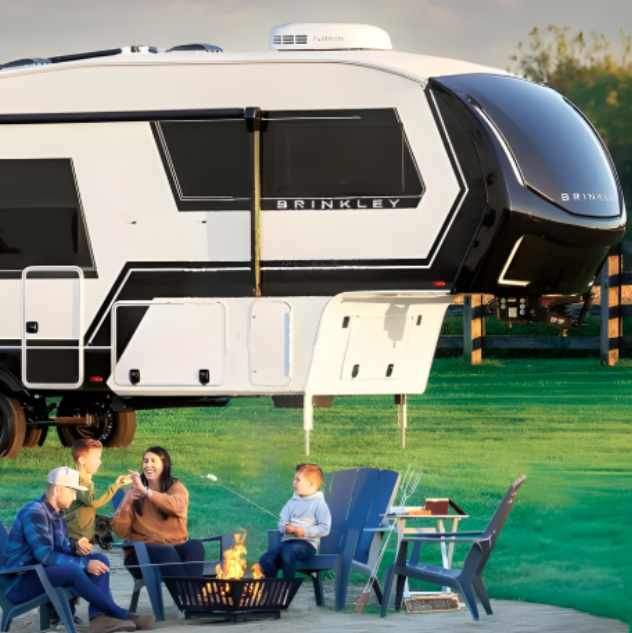 Family relaxing around a campfire beside a Brinkley fifth wheel RV, with outdoor chairs set up on a grassy campsite.