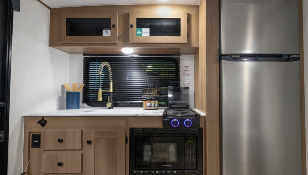 Compact travel trailer kitchen with sink, overhead cabinets, stovetop and oven with illuminated knobs, and full-size refrigerator to the right.