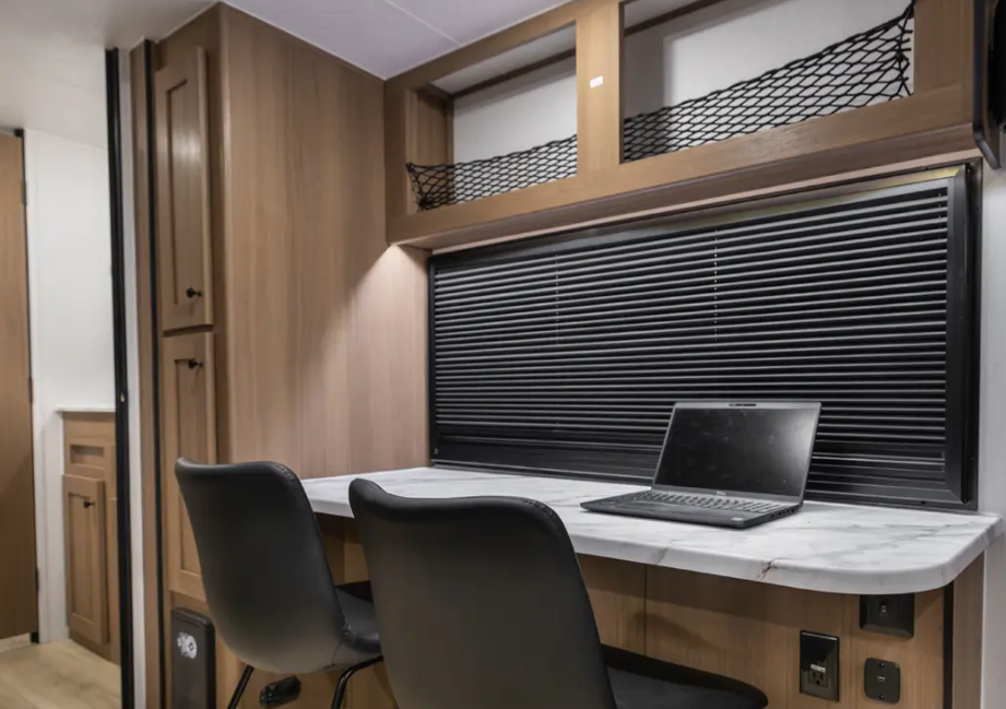 Travel trailer workspace with dual chairs, countertop desk, overhead storage with netting, and a window with closed blinds behind a laptop