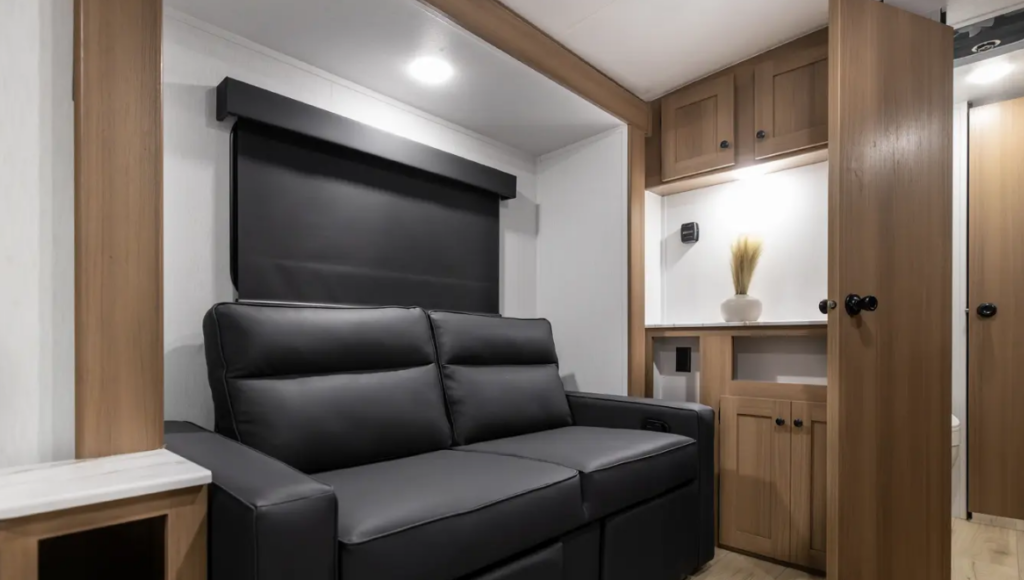 Travel trailer interior with black loveseat sofa beneath a window, adjacent cabinetry and storage, and hallway leading toward bathroom area