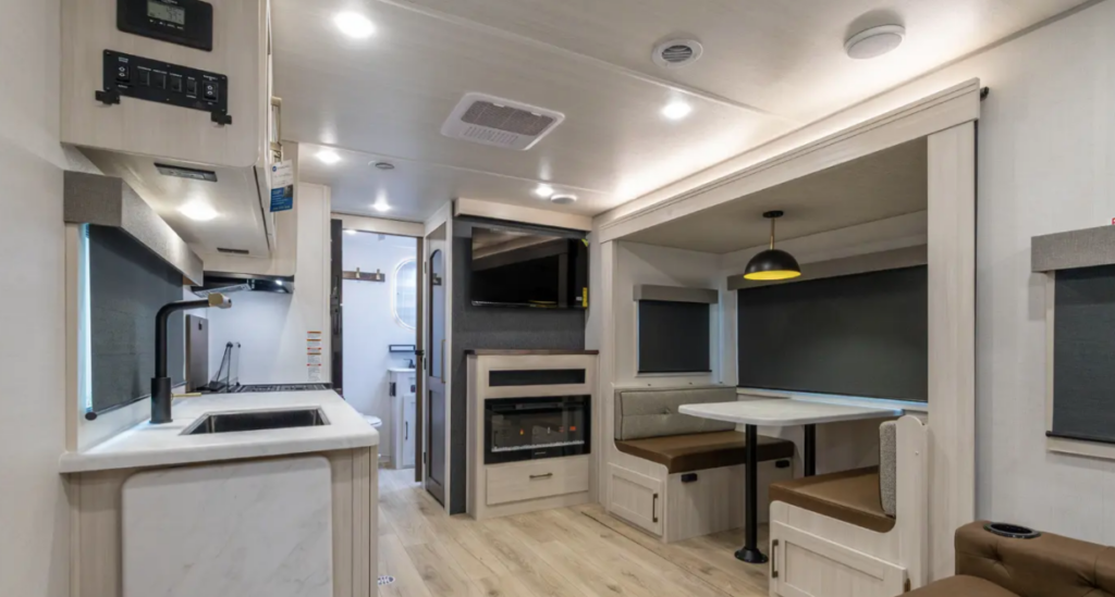 Travel trailer interior with kitchen sink and counter on the left, dinette booth on the right, and entertainment center with TV and fireplace in the center leading toward the bathroom