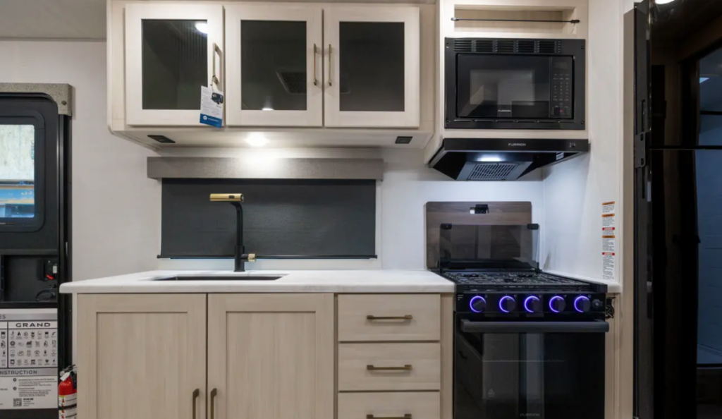 Travel trailer kitchen with sink, overhead cabinets, microwave, stove with illuminated knobs, and refrigerator to the side.
