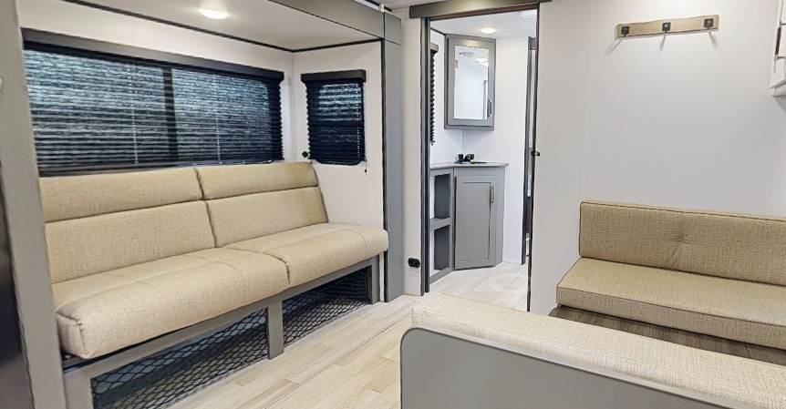 Travel trailer seating area with two bench-style sofas, windows with blinds, and a view into the bathroom vanity area.