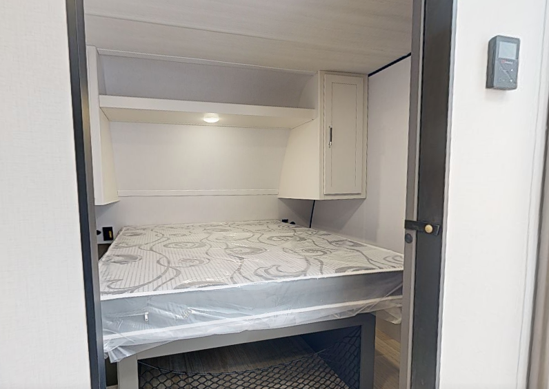 Travel trailer bedroom with queen bed, overhead shelf, side cabinet, and under-bed storage visible through a sliding doorway.