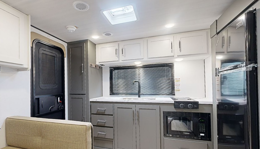 Travel trailer kitchen with gray cabinetry, sink under a window with blinds, microwave and stove below, and refrigerator on the right.