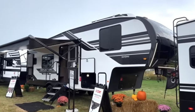 Reflection fifth wheel RV on display outdoors with awning extended, entry steps down, and seasonal decorations including pumpkins and flowers.