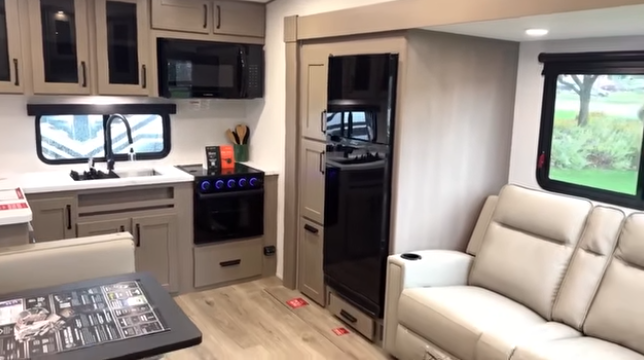 Travel trailer interior showing kitchen with sink, stove, and microwave, adjacent refrigerator, and a light-colored reclining sofa by the window.