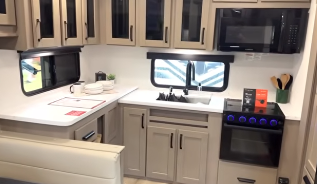Travel trailer kitchen with L-shaped countertop, sink beneath a window, overhead cabinets, microwave, and stove with illuminated knobs.