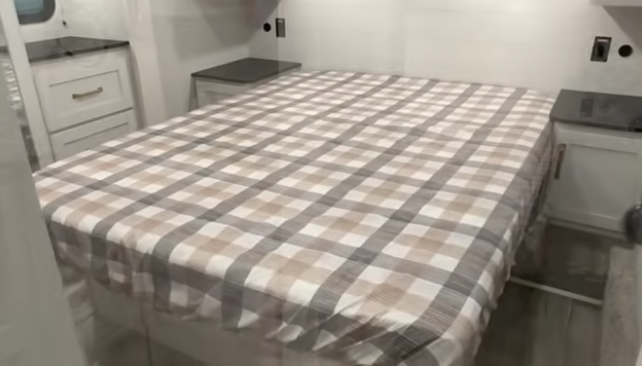 Travel trailer bedroom with a queen bed featuring a plaid bedspread, flanked by small nightstands and overhead storage.