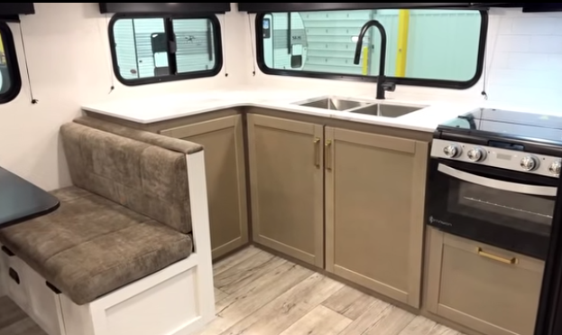 Travel trailer kitchen with L-shaped countertop, double sink under windows, stove and oven, and adjacent booth-style dinette seating.