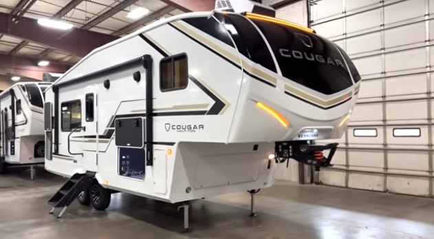Cougar fifth wheel RV displayed indoors with LED accent lighting, front hitch visible, entry steps down, and side storage compartments open.