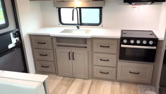 Travel trailer kitchen with wide countertop, sink under a window, multiple drawers and cabinets, and a stove and oven on the right.