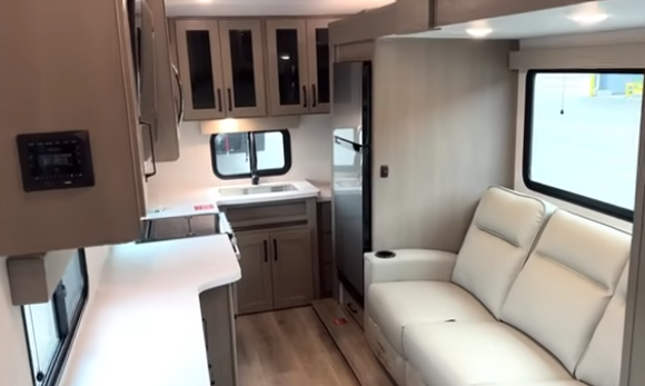 Compact travel trailer interior with kitchen counter and sink, overhead cabinets, refrigerator, and a light-colored sofa along the wall.