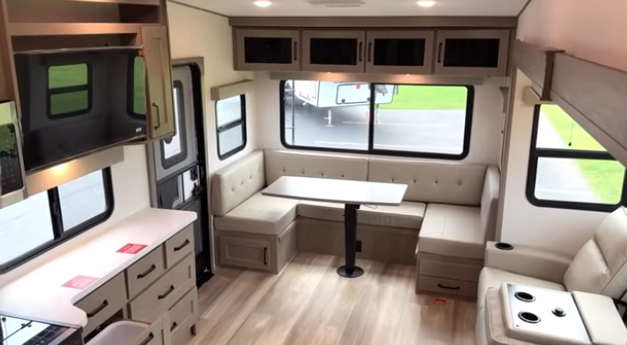 Travel trailer interior with U-shaped dinette under large windows, overhead cabinets, and adjacent seating and storage cabinetry.