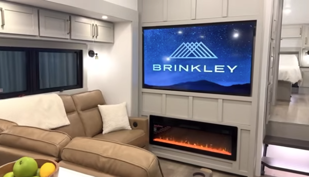Travel trailer living area with leather sofa, large TV displaying Brinkley logo, and electric fireplace below, with bedroom visible in the background.