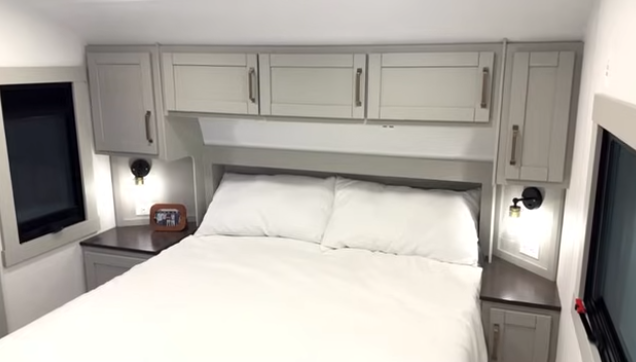 Travel trailer bedroom with a queen bed, overhead cabinets, bedside tables with reading lights, and windows on both sides.