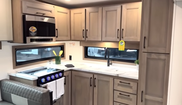 Travel trailer kitchen with L-shaped countertop, gray cabinetry, sink beneath a window, microwave above the stove, and oven with illuminated knobs.