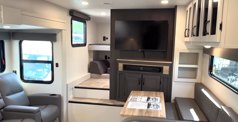 Travel trailer interior with entertainment center and TV, dinette seating, reclining chair, and a raised rear sleeping area with steps.