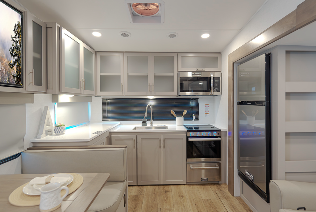 Travel trailer kitchen with white cabinetry, sink under a window with blinds, stove and oven, microwave above, and refrigerator to the right next to a booth dinette.
