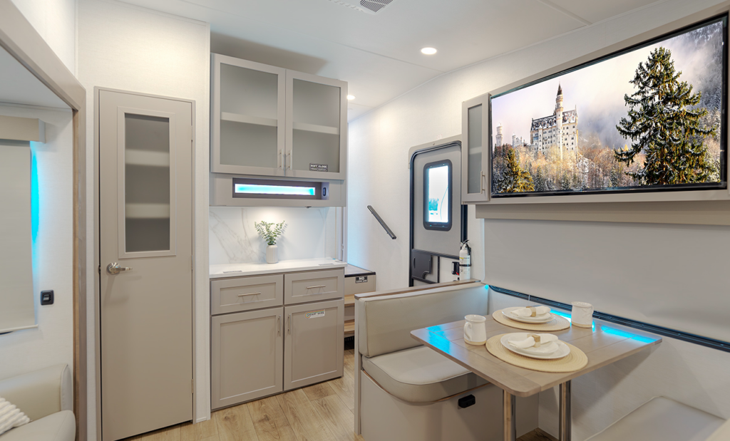 Travel trailer interior with booth dinette, overhead cabinetry, pantry storage, entry door, and wall-mounted TV above accent lighting.
