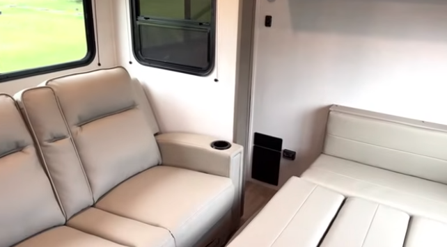 Travel trailer seating area with a light-colored sofa featuring a cup holder, next to a fold-down bed or convertible seating under a window.