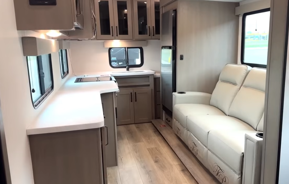 Compact travel trailer interior with extended kitchen counter, sink and cabinetry, refrigerator, and a light-colored sofa along the wall.