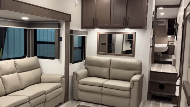 Travel trailer living area with two light-colored sofas, overhead cabinets, and steps leading up to a rear bedroom.
