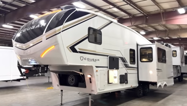 Keystone Cougar half-ton fifth wheel RV displayed indoors with slide-out extended, front hitch elevated, and LED accent lighting on the front cap.