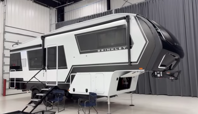 Brinkley fifth wheel RV displayed indoors with slide-out extended, entry steps down, and outdoor chairs set up beneath the awning.