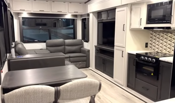 Travel trailer interior with dinette table and chairs, kitchen with stove and microwave, entertainment center, and a rear sofa beneath large windows.