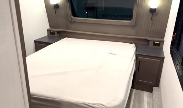 Travel trailer bedroom with a queen bed, side nightstands, reading lights, and a window above the headboard.