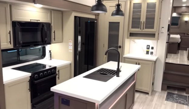 Travel trailer kitchen with center island and sink, pendant lighting, stove and microwave, refrigerator, and cabinetry, with steps leading to a rear bedroom.