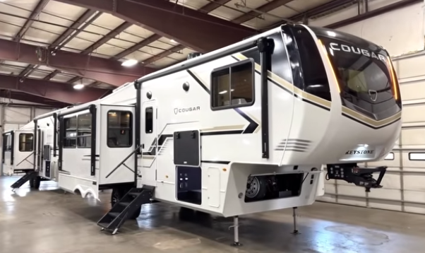 Keystone Cougar fifth wheel RV displayed indoors with slide-outs extended, entry steps down, and front hitch elevated.