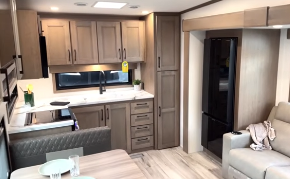Travel trailer interior with kitchen featuring wood cabinetry, sink under a window, tall pantry and refrigerator, and a sofa next to the living area.