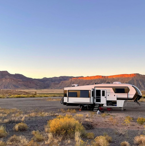 Fifth wheel RV parked in a desert landscape at sunset with mountains in the background and slide-out extended.