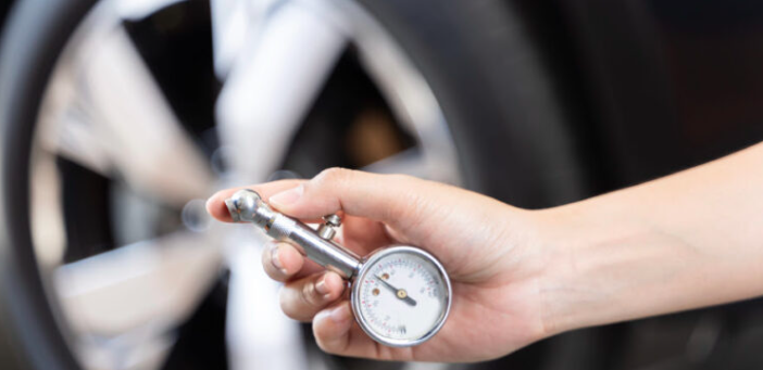 Hand holding a tire pressure gauge in front of a vehicle wheel, measuring air pressure.