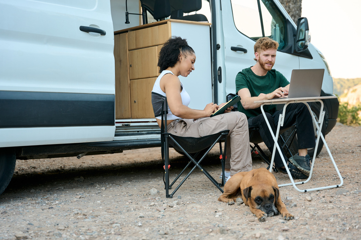 Young multi-ethnic couple working remotely using laptop and tablet near their camper van with their dog