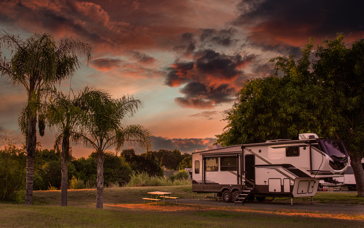 A recreational vehicle is parked amidst lush greenery under a dramatic, colorful sky at sunset. Palm trees sway gently in the foreground, adding to the tranquil atmosphere of the campsite. The vibrant hues of the sunset contrast beautifully with the darkening sky, creating a serene and inviting scene.