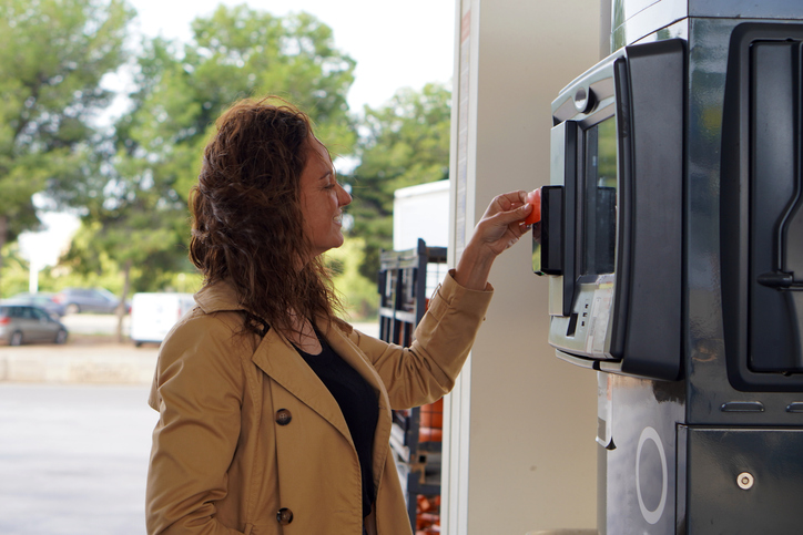A middle-aged woman with curly hair and a brown trench coat, smiling as she pays with her card at a gas station.