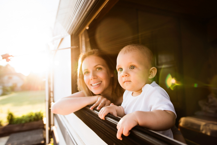 Smiling woman and baby leaning out of a travel trailer window, looking outside in warm sunlight.