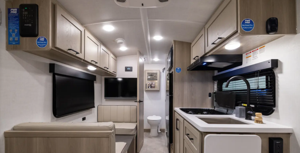 Compact travel trailer interior with kitchenette, booth dinette, overhead cabinets, TV, and rear bathroom visible through the hallway.