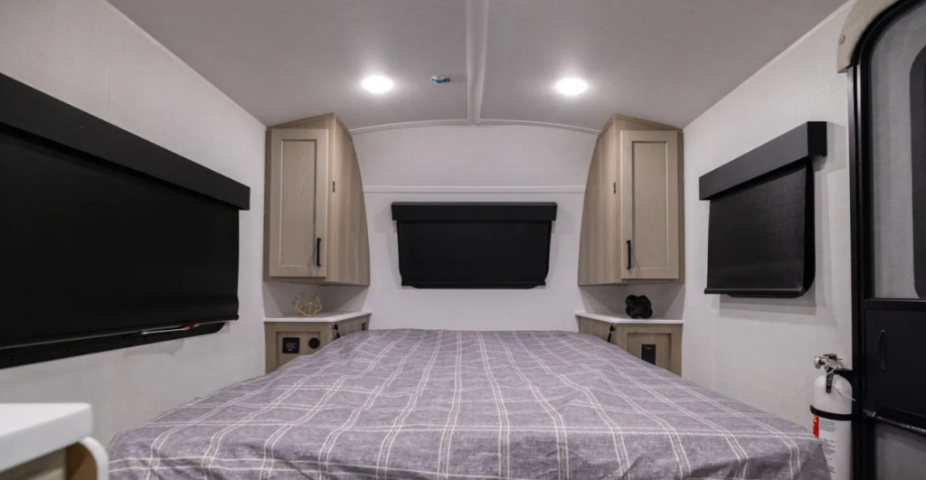 Compact RV bedroom with a bed centered between bedside cabinets, blackout window shades, and a wall-mounted TV