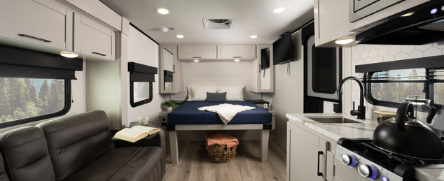 Compact RV interior with kitchenette, black leather sofa, mounted TV, and rear bed with storage basket underneath.