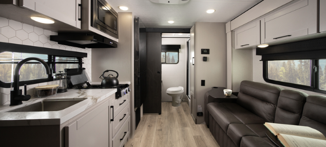 Compact RV interior with modern kitchen, black leather sofa, overhead cabinets, and hallway leading to a rear bathroom.