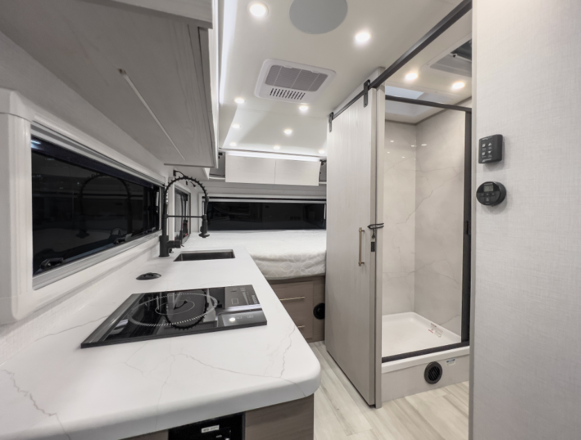 Compact RV interior with modern kitchenette, induction cooktop, sink, sliding bathroom door, shower, and rear sleeping area.