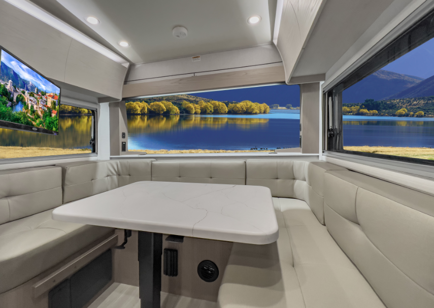 RV dinette area with wraparound seating, large windows overlooking a lake and mountains, and a wall-mounted TV.