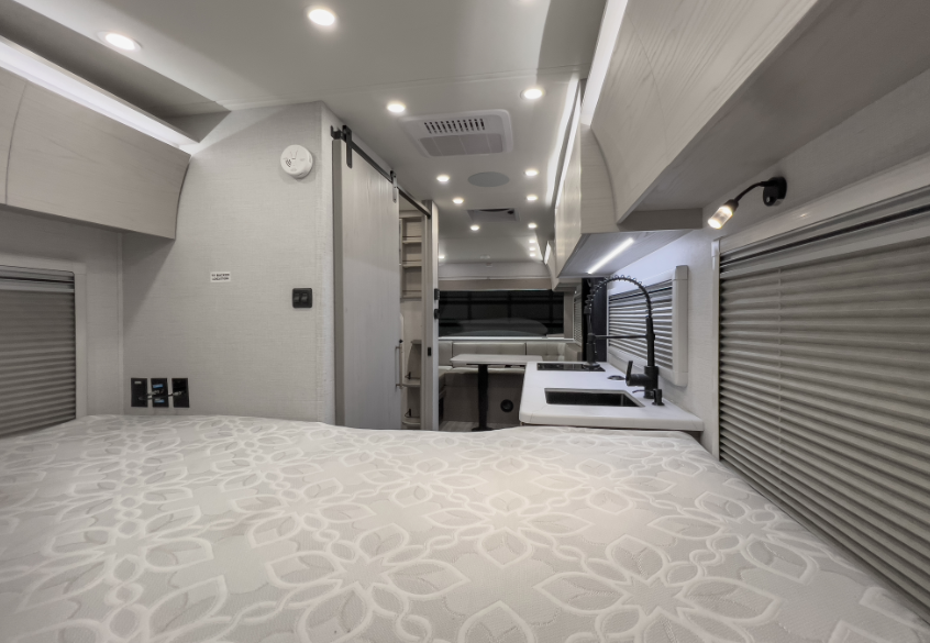 Compact RV interior viewed from the bed, featuring overhead storage, kitchenette with sink, window shades, and rear seating area.
