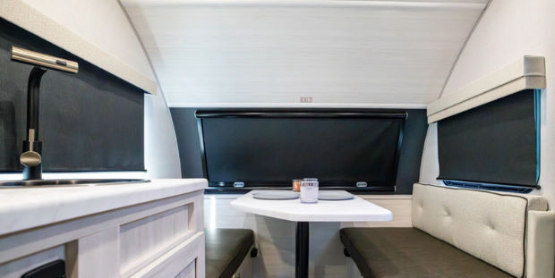 Compact RV dinette area with bench seating, small table, kitchenette counter with sink, and blackout window shades.